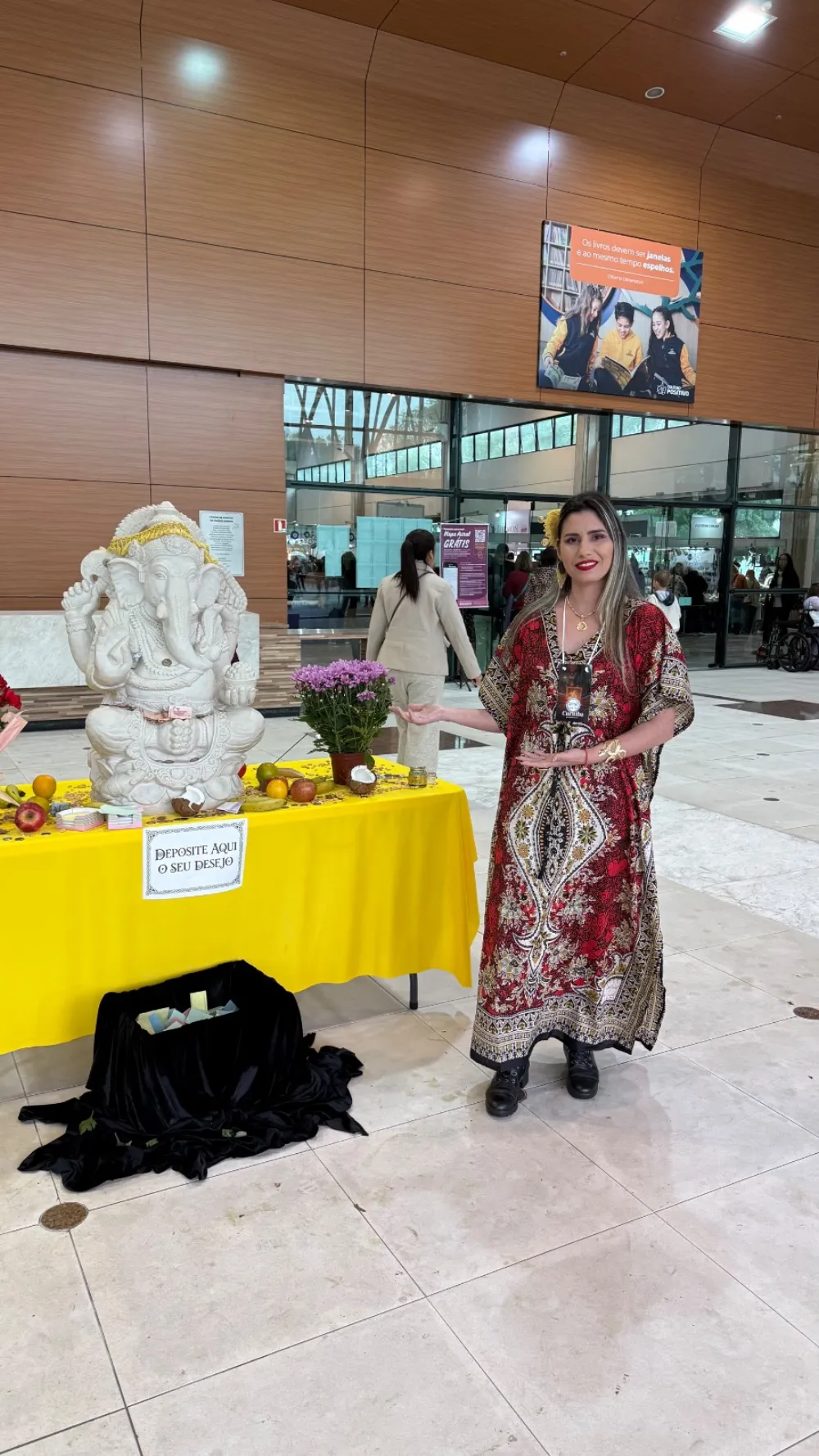 Sarah Sensitiva ao lado de uma estátua de Ganesha em uma feira, com a mesa dos desejos