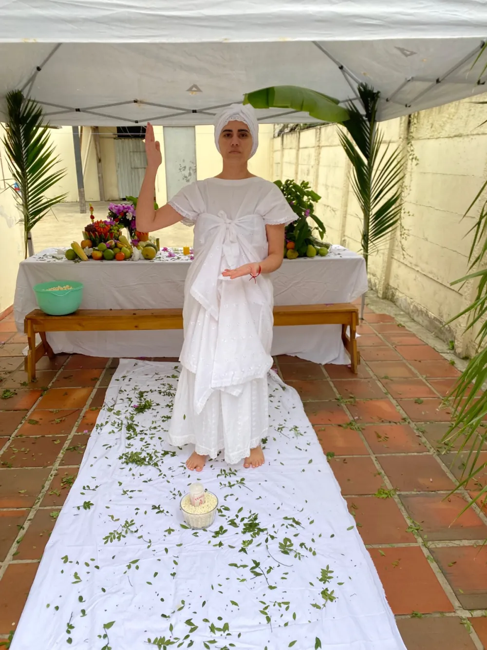 Sarah vestida de branco em um ritual, com mesa de frutas e folhas no chão