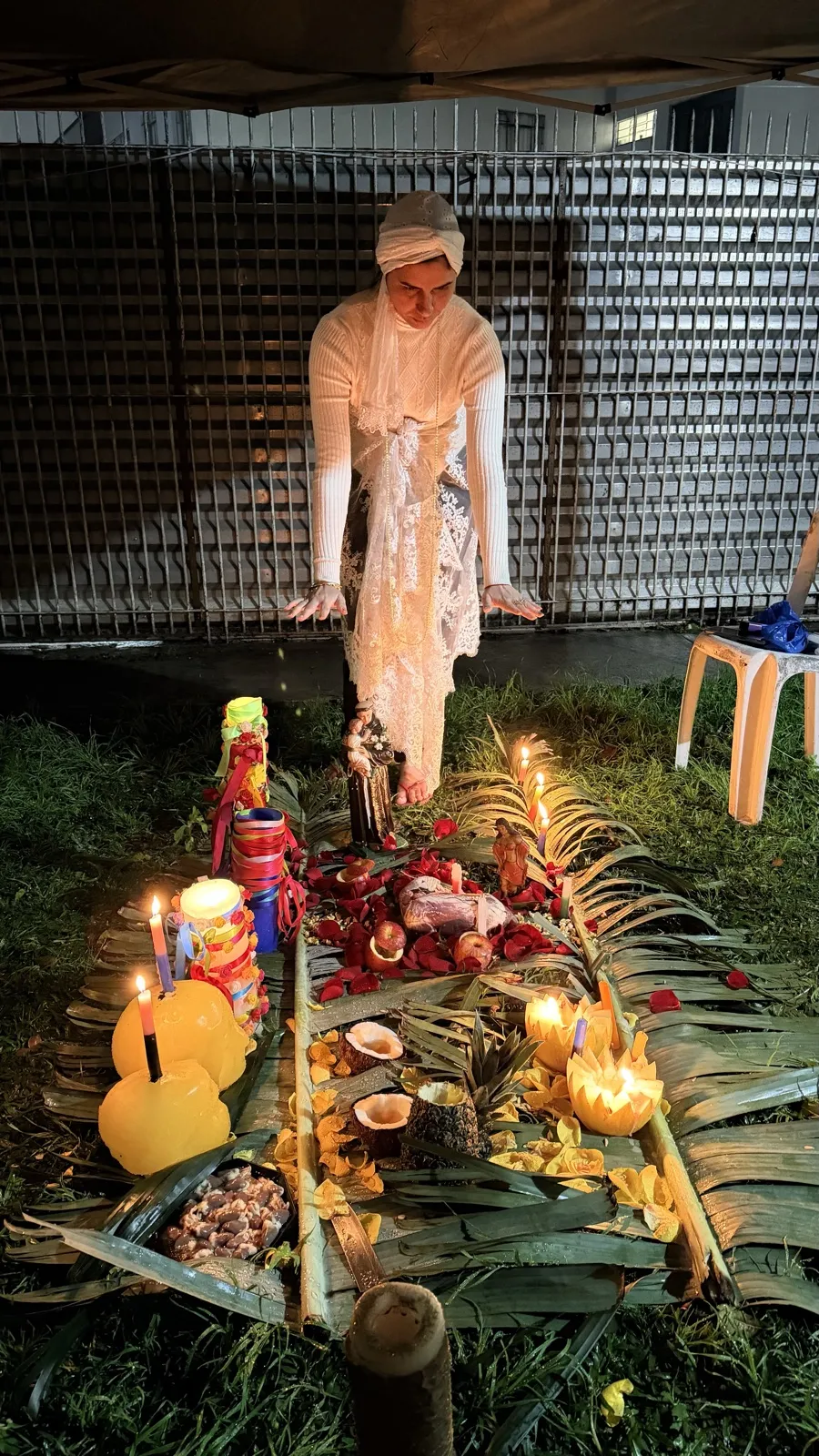 Sarah de branco ao fundo de uma oferenda no gramado, com folhas de palmeira, frutas e velas à noite